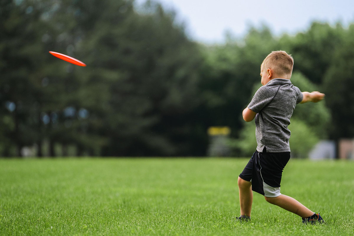 A young boy wearing a gray shirt and black shorts throws a red frisbee across a grassy field, with trees blurred in the background.