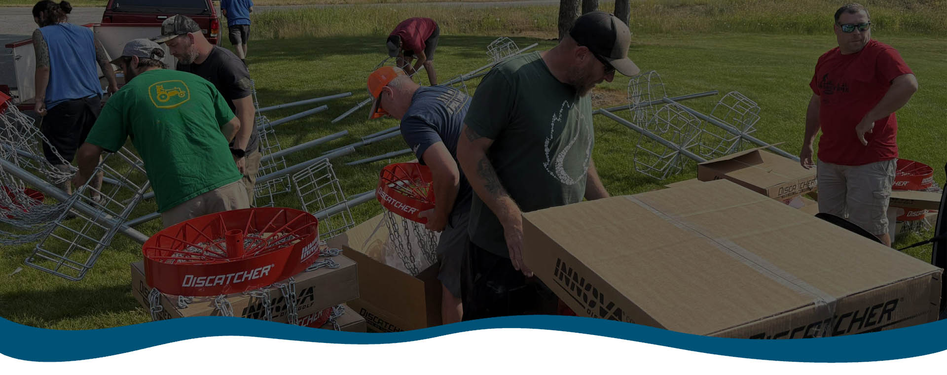 A group of people assemble metal disc golf baskets and unpack boxes outdoors on a grassy area under sunny weather.