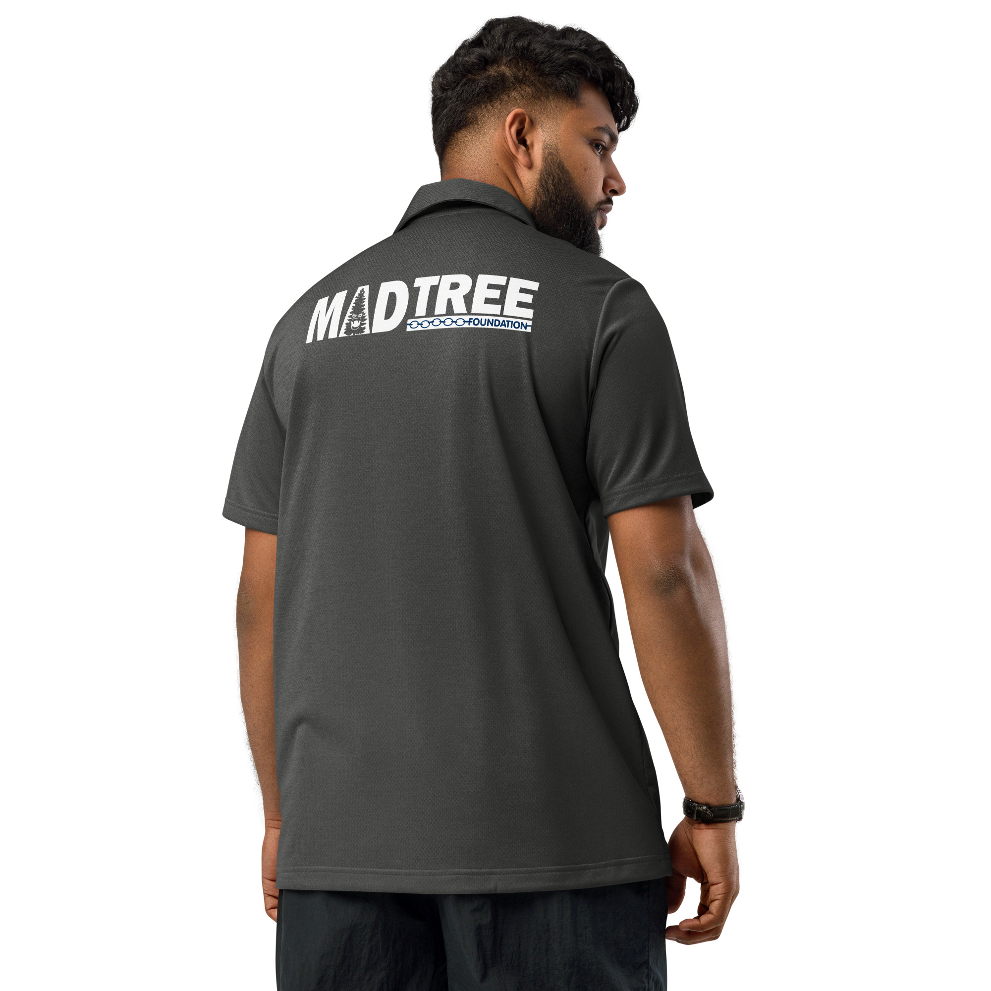 A man with a beard, short hair, and black pants stands facing away, wearing a dark gray adidas space-dyed polo with MADTREE and a white tree logo printed on the back.