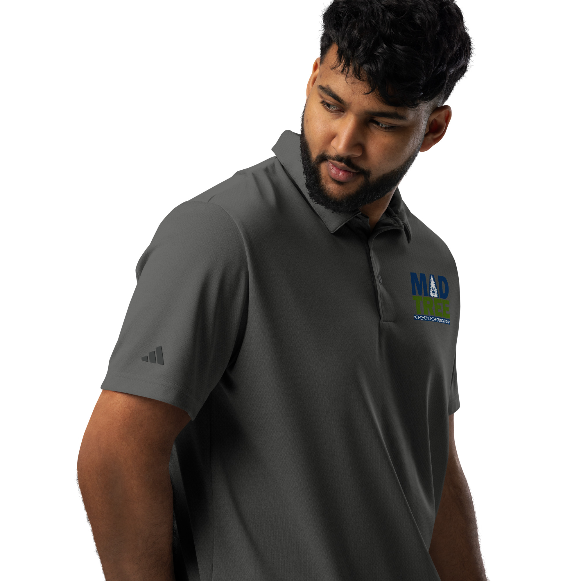 A man with short dark hair and a beard wears an adidas space-dyed polo featuring the MAD TREE logo on the chest and Adidas logo on the sleeve, looking down to his left against a white background.