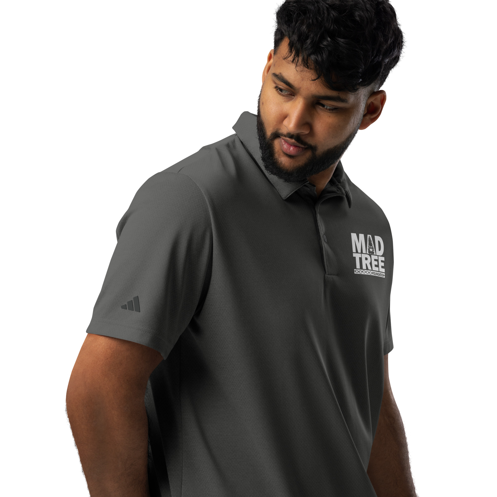 A man with curly hair and a beard wears an adidas space-dyed polo in dark gray with MAD TREE branding and a small Adidas logo on the sleeve, looking down to his left against a plain white background.