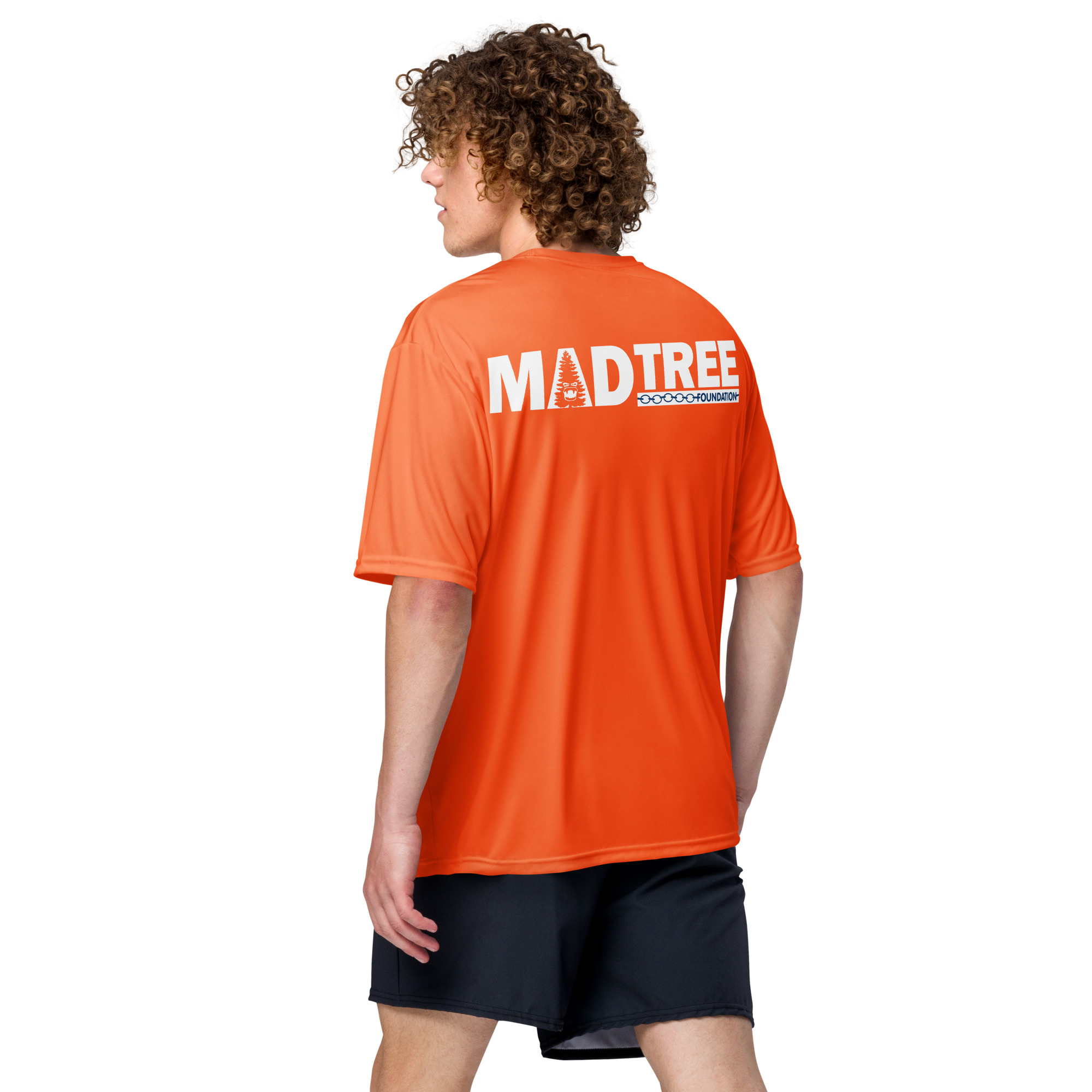 A person with curly hair wearing an orange Unisex performance crew neck t-shirt and black shorts stands facing away, looking over their left shoulder against a plain white background.