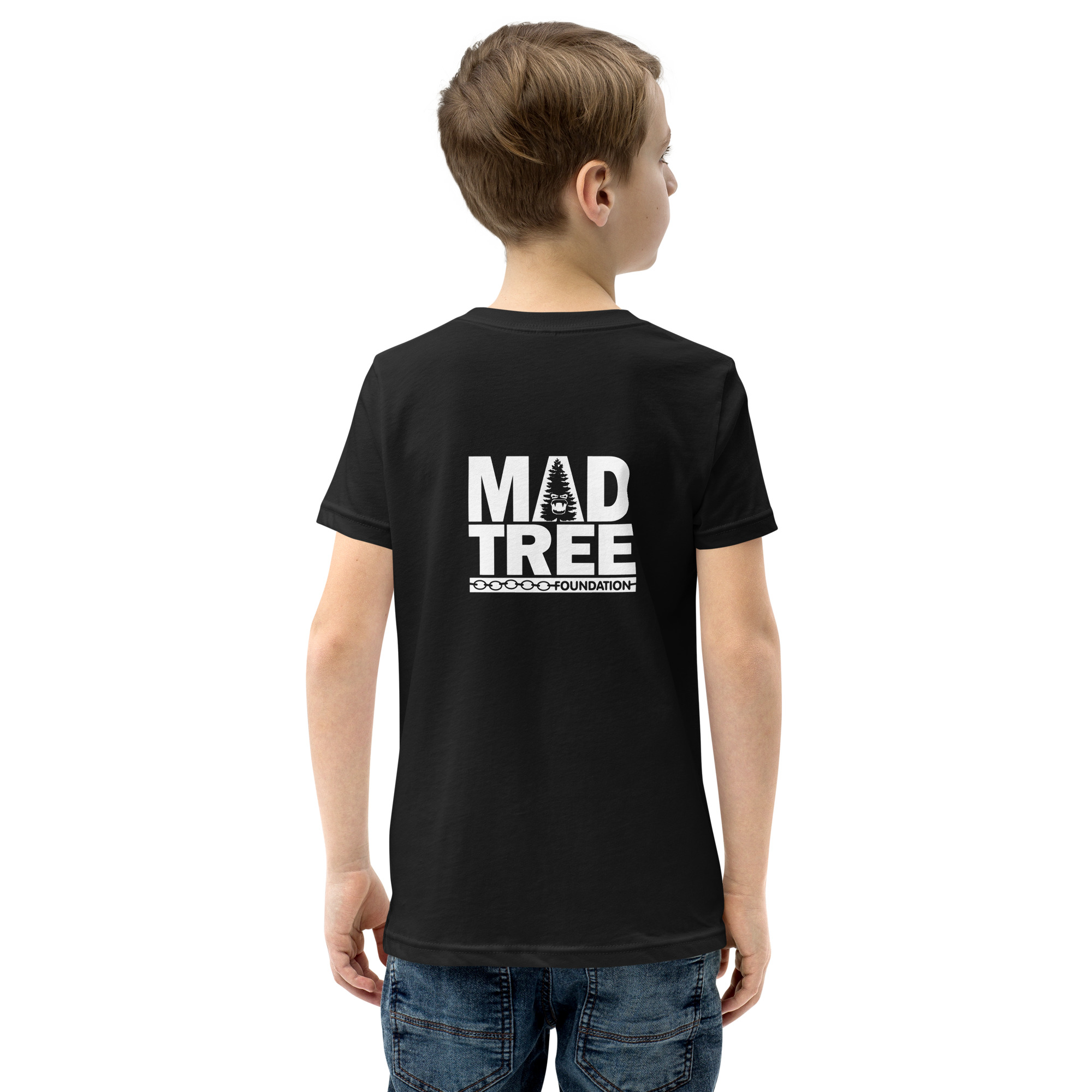A boy wearing the Youth Short Sleeve T-Shirt - Mad Tree, featuring "MAD TREE FOUNDATION" and a white tree graphic on the back, stands in jeans facing away from the camera.